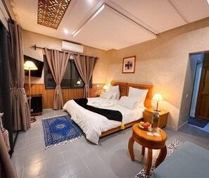 A hotel room with a bed, bedside lamps, a small table with fruit, a blue rug, and an air conditioning unit. - Atlas Front (Sidi Abdallah Ghiat)