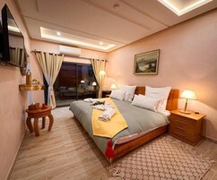 Una habitación de hotel con cama, lámparas de noche, una mesita, televisor y ventana con cortinas.