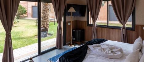 A hotel room with a large bed, a flat-screen TV, a small table with a bottle and a plate, and a view of a garden and building outside.