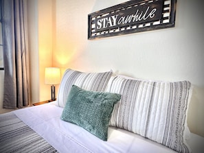 A neatly made bed with striped bedding and a green pillow, a wall-mounted sign saying "STAY awhile", and a lamp on a bedside table.