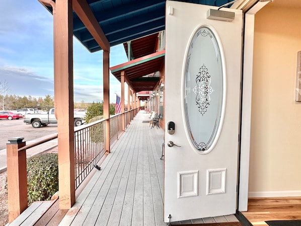 A wooden deck with a covered porch, a white door with decorative glass, and a view of parked cars and trees in the background.