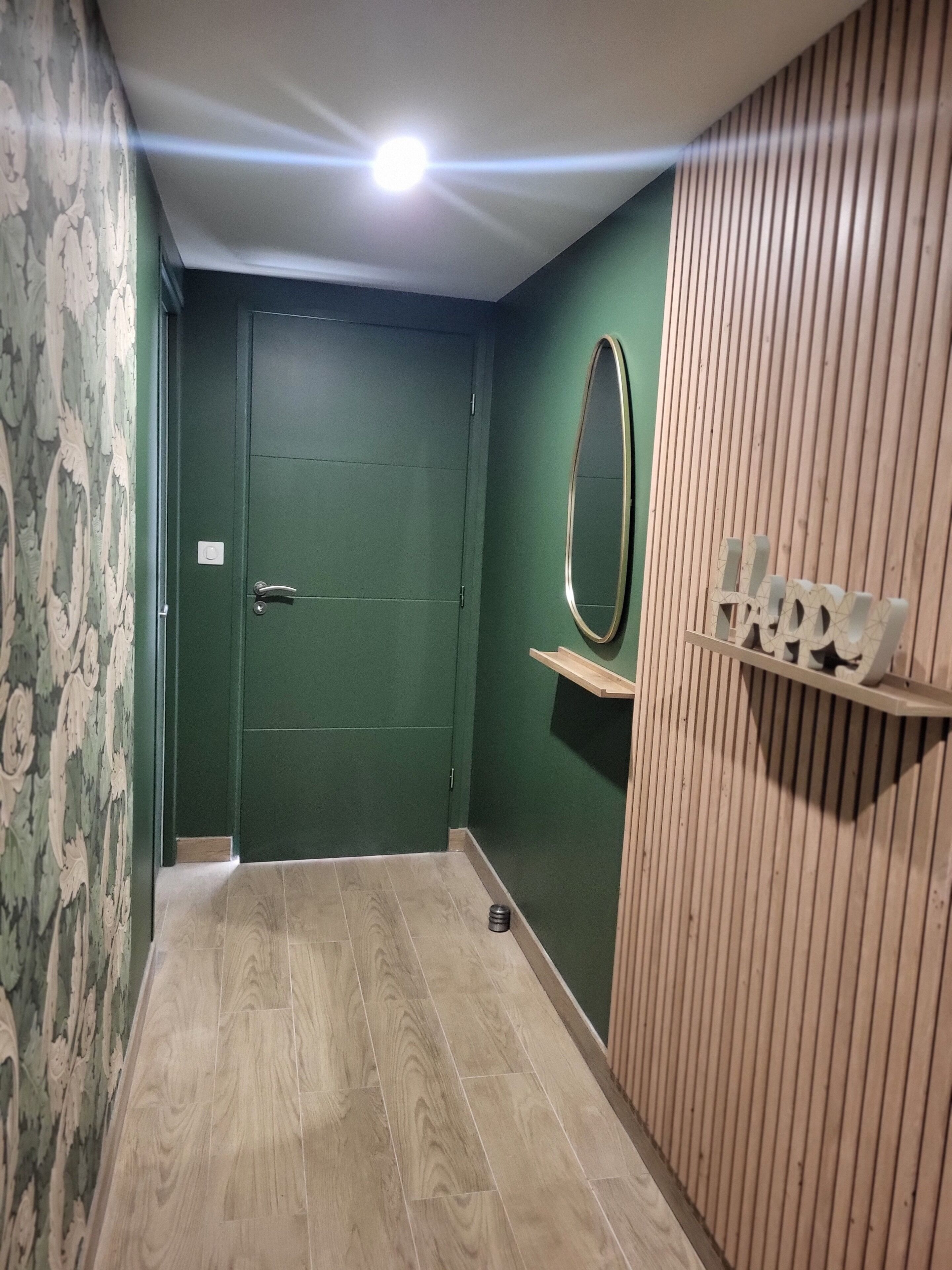 Un couloir avec une porte verte, un plancher en bois, un miroir et un panneau indiquant le mot « Heureux » sur le mur.