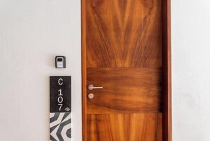A wooden door with a metal handle and a keyhole, a doorbell, and a black and white sign with the number "C107" on a white wall. - C107 CPL, Relaxing Studio Garden Pool (Tulum)