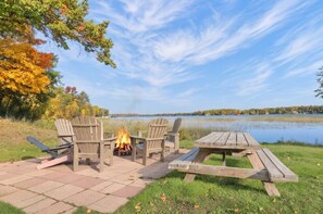 A lakeside outdoor seating area with a fire pit, chairs, and a table.