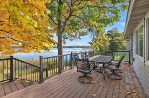 A wooden deck with a table and chairs, overlooking a lake with autumn foliage.