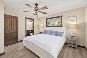 A bedroom with a large bed, a ceiling fan, a window with a view, and a wooden door.