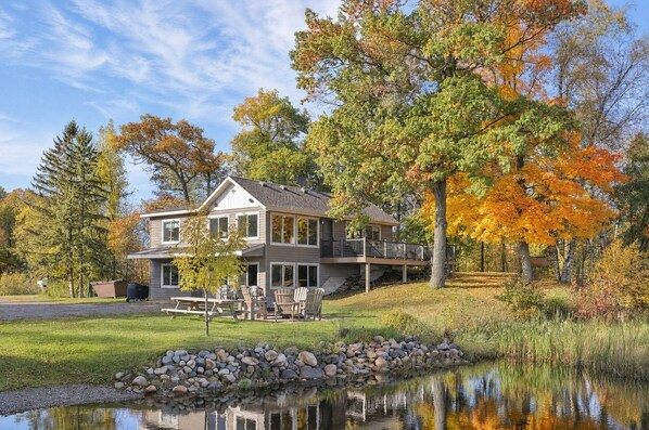 A two-story house with a deck, a pond in the foreground, and trees with autumn foliage in the background.