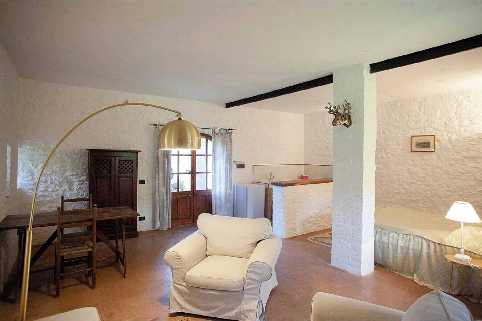A room with a wooden dining table, a white armchair, a sofa, a lamp, a kitchen area, and a window with curtains.
