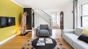 Una sala de estar moderna con un sofá gris, una mesa de centro negra con revistas, piso de madera, una escalera y un ventanal grande.