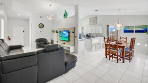 A modern living room with a grey sofa, a TV, and a dining area with a wooden table and chairs. - Waterfront Oasis – 4BR Pool & Spa (Kissimmee)
