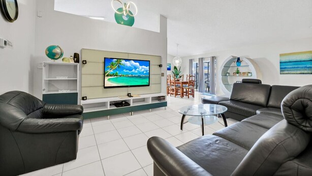 A modern living room with a flat-screen TV, leather sofas, a glass coffee table, and a dining area with wooden chairs. - Waterfront Oasis – 4BR Pool & Spa (Kissimmee)