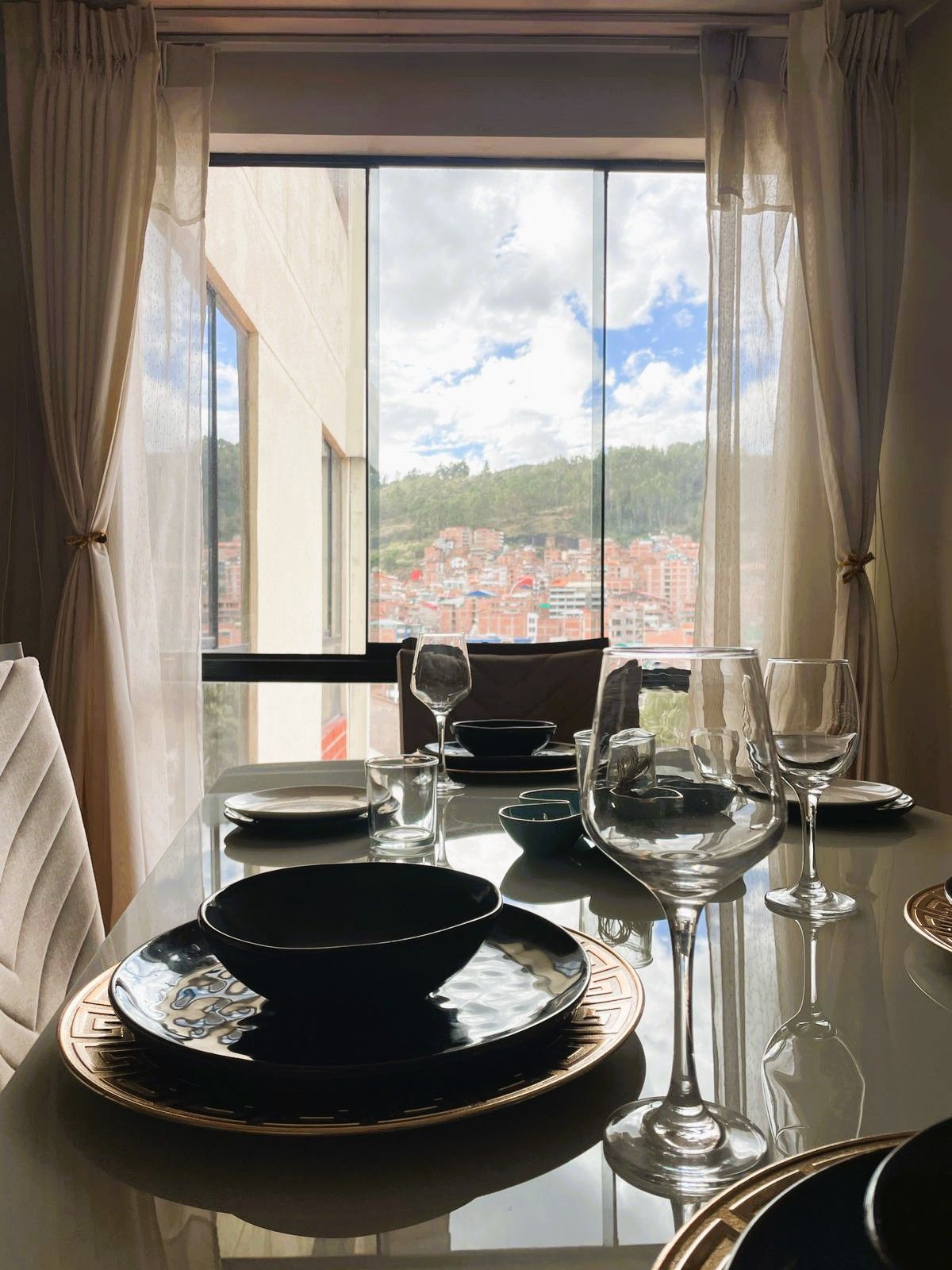 Une table à manger dressée avec des assiettes, des bols et des verres à vin, donnant sur un paysage urbain à travers une fenêtre.