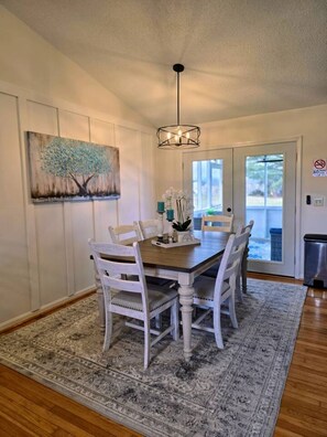 A dining area with a wooden table and white chairs, a chandelier, a painting of a tree, and a view through a glass door. - Peaceful Valley Hideaway—a roomy & fun, family-friendly home in Jonesborough, TN (Jonesborough)