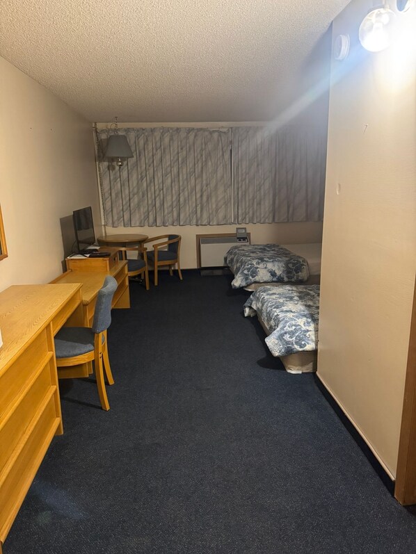 Ett kompakt hotellrum med ett skrivbord, en stol, två enkelsängar, ett litet bord och en lampa.
