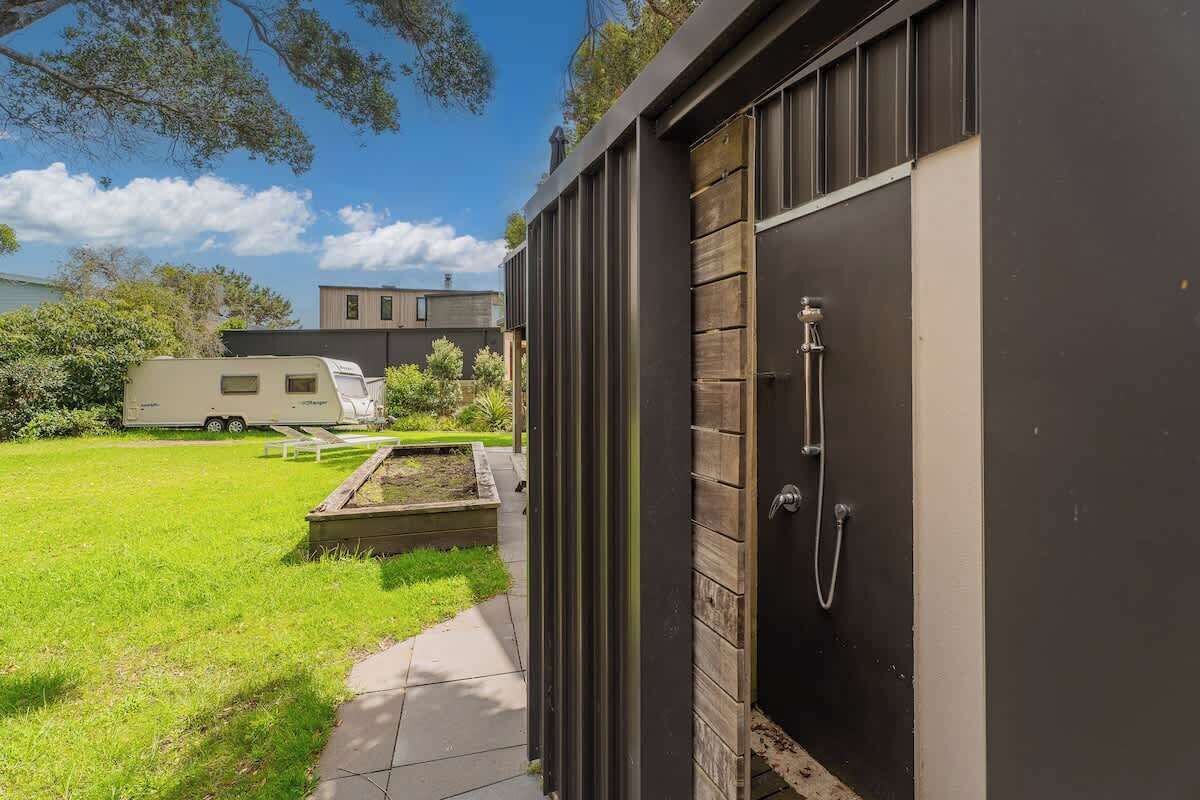 Un área de ducha exterior moderna con una puerta de madera, un sendero que conduce a una zona cubierta de césped y una caravana al fondo.