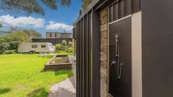 Un área de ducha exterior moderna con una puerta de madera, un sendero que conduce a una zona cubierta de césped y una caravana al fondo.