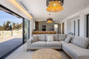 A modern living room with a large sectional sofa, a round wooden coffee table, and a kitchen in the background. - Villa Maninee-Bespoke Villa-Pool -BBQ-Seaview (Chania)