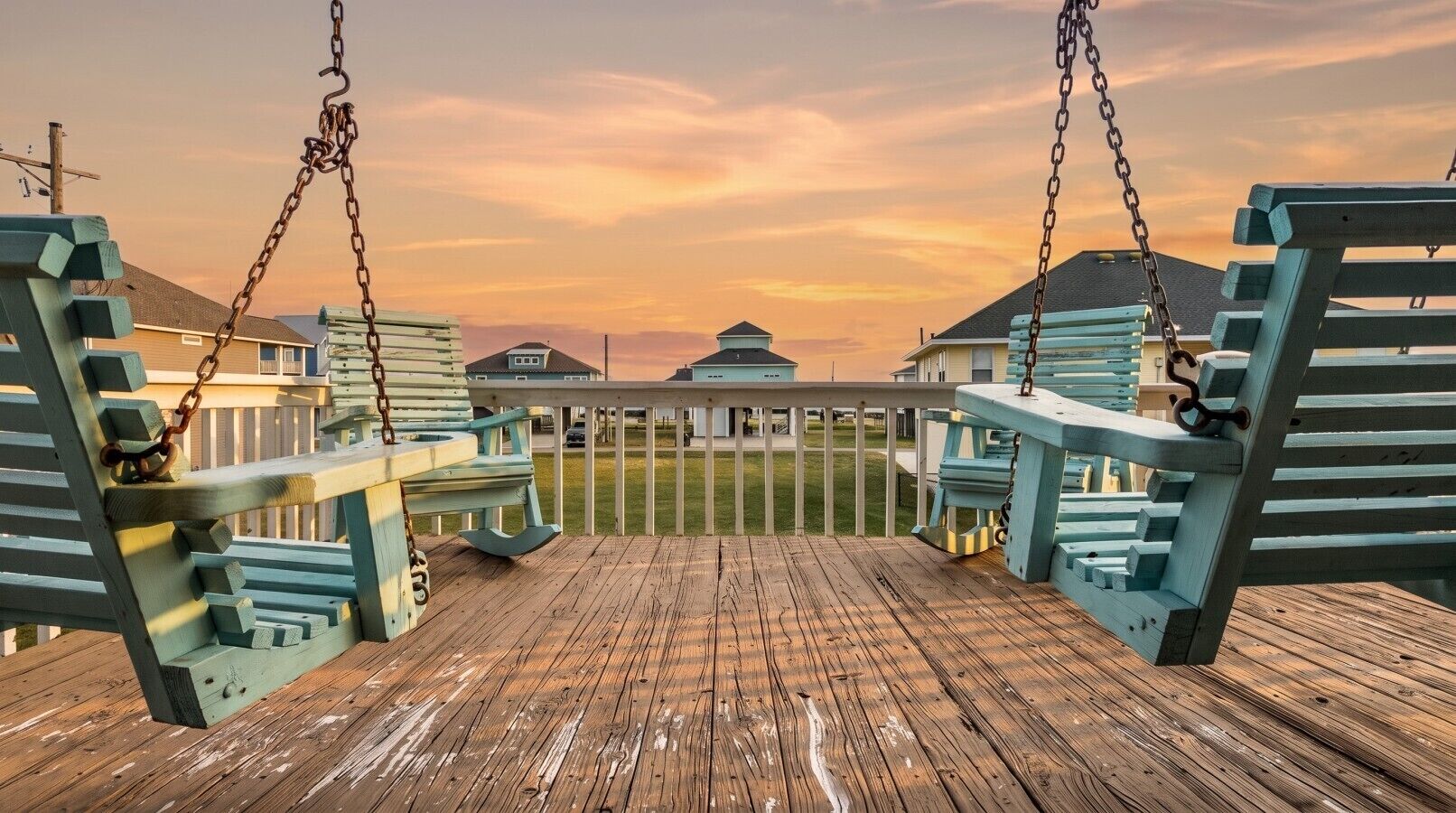 A wooden deck with a swing set, a white fence, and houses in the background.