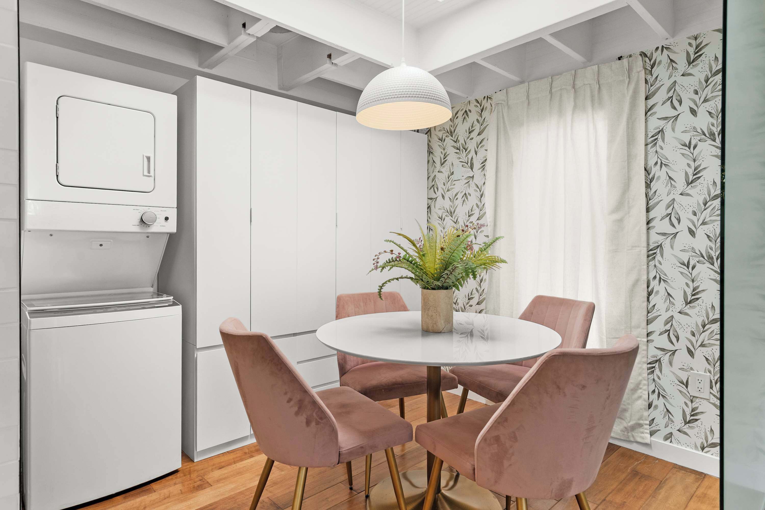 A modern kitchen with white cabinets, a built-in oven, a round dining table with pink chairs, and a hanging pendant light.