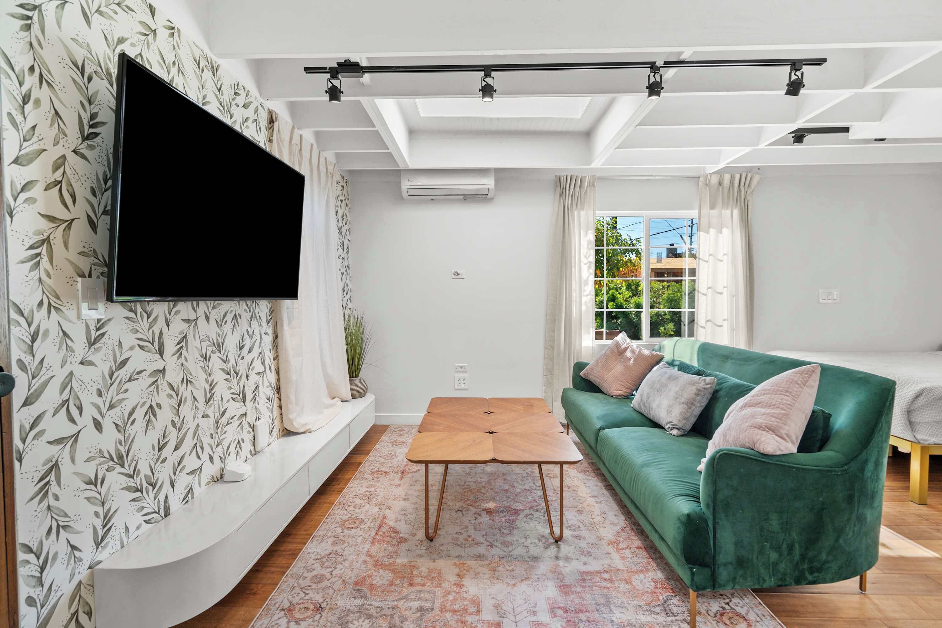 A modern living room with a flat-screen TV mounted on the wall, a green sofa with cushions, a wooden coffee table, and a patterned rug.
