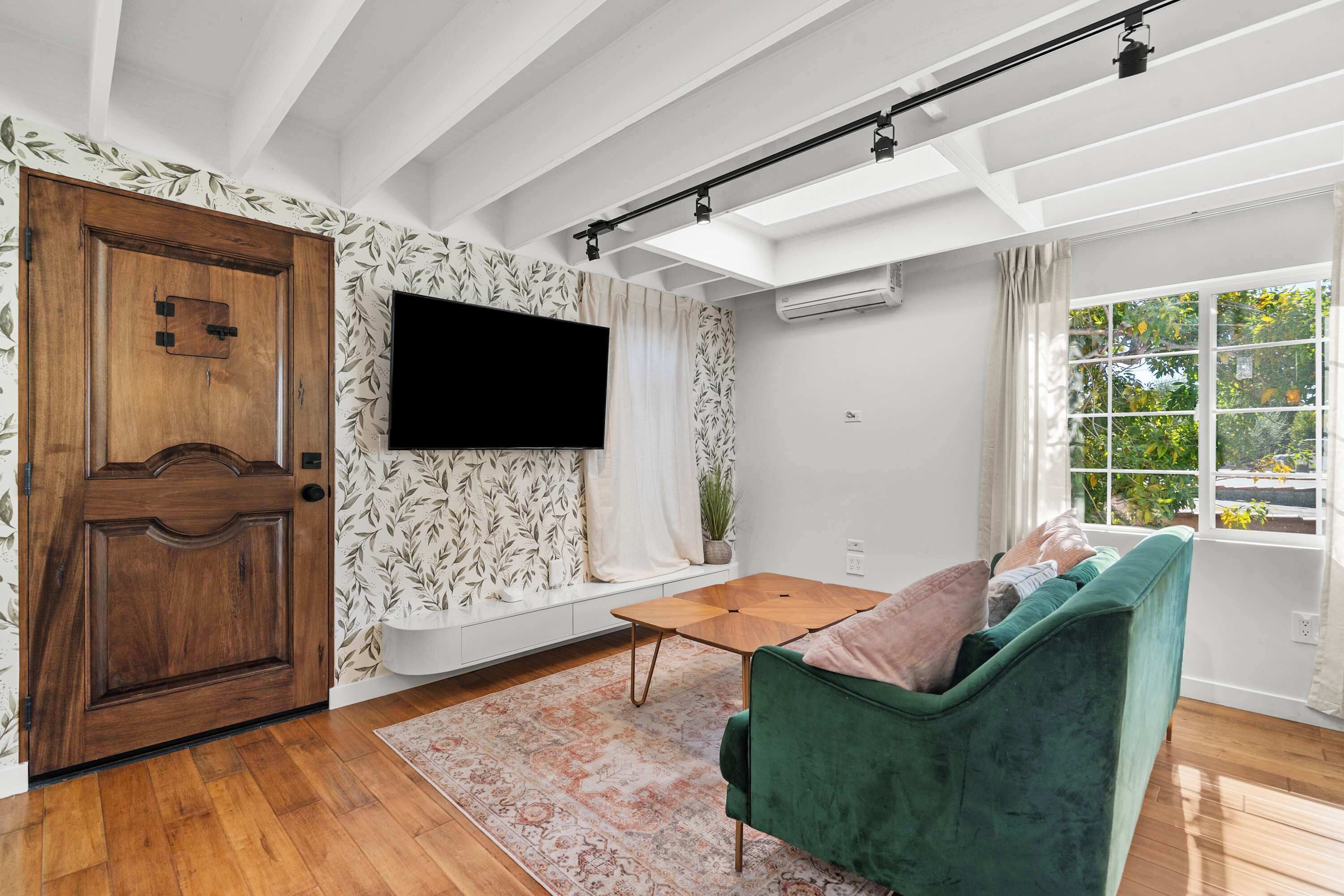 Ein Wohnzimmer mit einem grünen Sofa, Holzboden, einem Wand-Fernseher und einem Fenster mit Blick auf Grünflächen.
