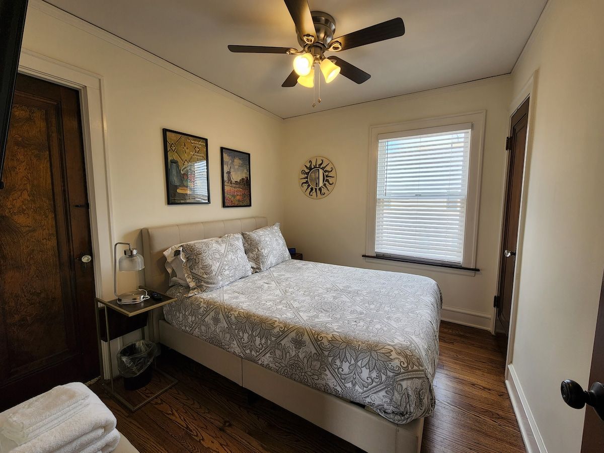 Une chambre à coucher comprenant un lit, un ventilateur de plafond, une table de chevet, une lampe, une fenêtre avec des stores et une porte.