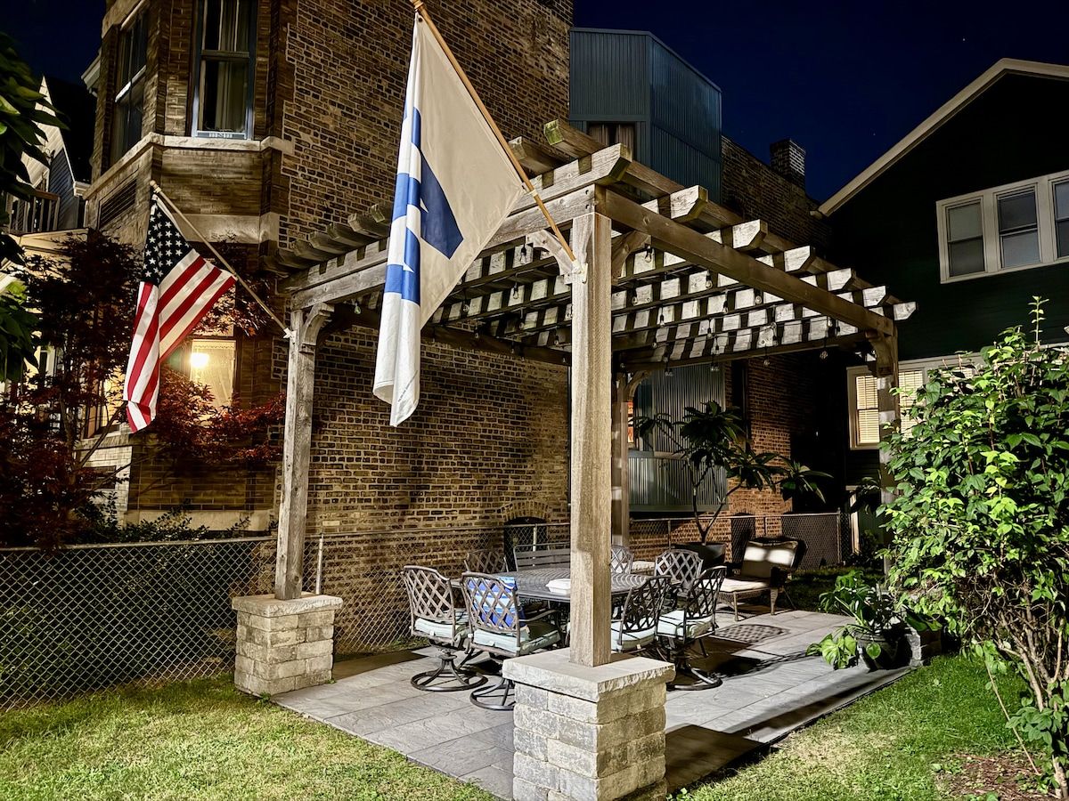 Ein überdachter Patio-Bereich mit Gartenmöbeln, einer Flagge und einem Backsteinhaus im Hintergrund.