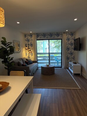 A modern living room with a sofa, a dining table, a large window with a view, and a flat-screen TV mounted on the wall. - Beautifully Renovated condo in Hilton Head Island- 5 min walk to private beach (Hilton Head Island)
