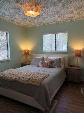 Une chambre à coucher comprenant un lit, deux tables de chevet, une commode, un plafond à motifs et deux fenêtres équipées de stores.