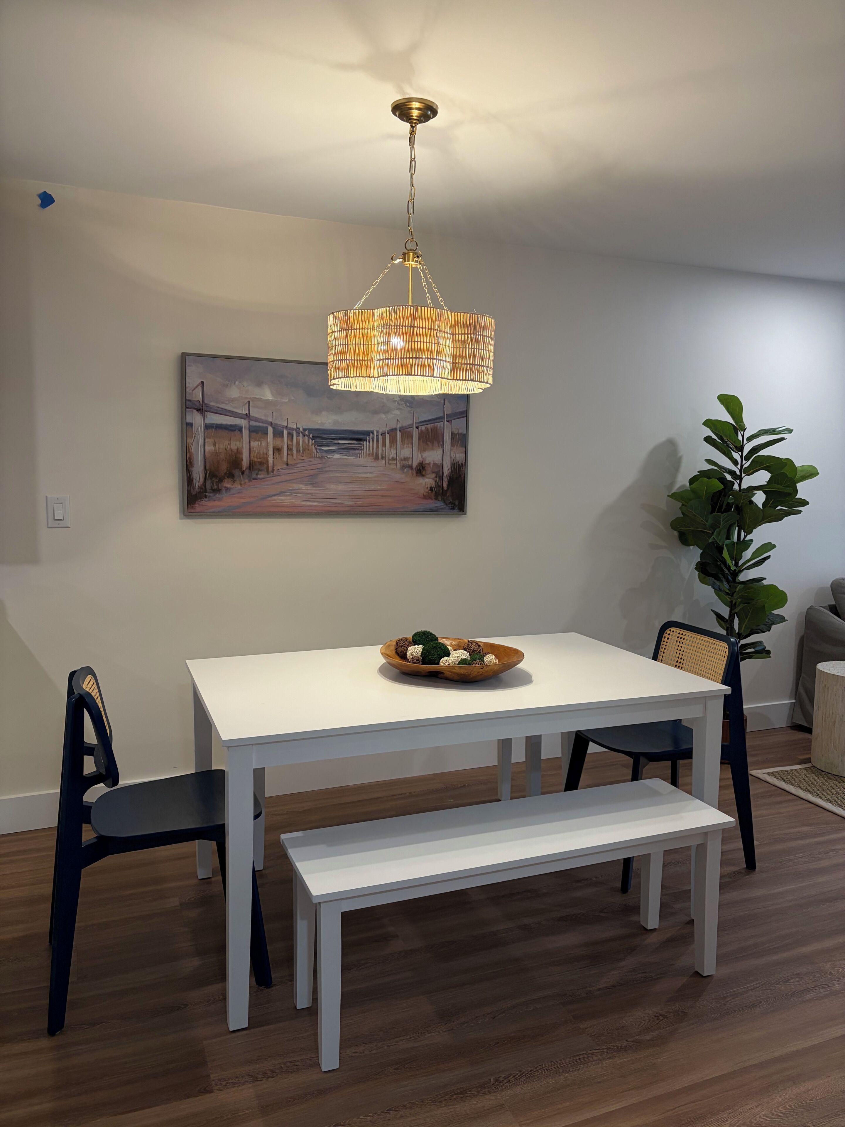 Un coin repas avec une table blanche, deux chaises, un banc, un lustre suspendu en osier, un tableau au mur et une plante en pot.