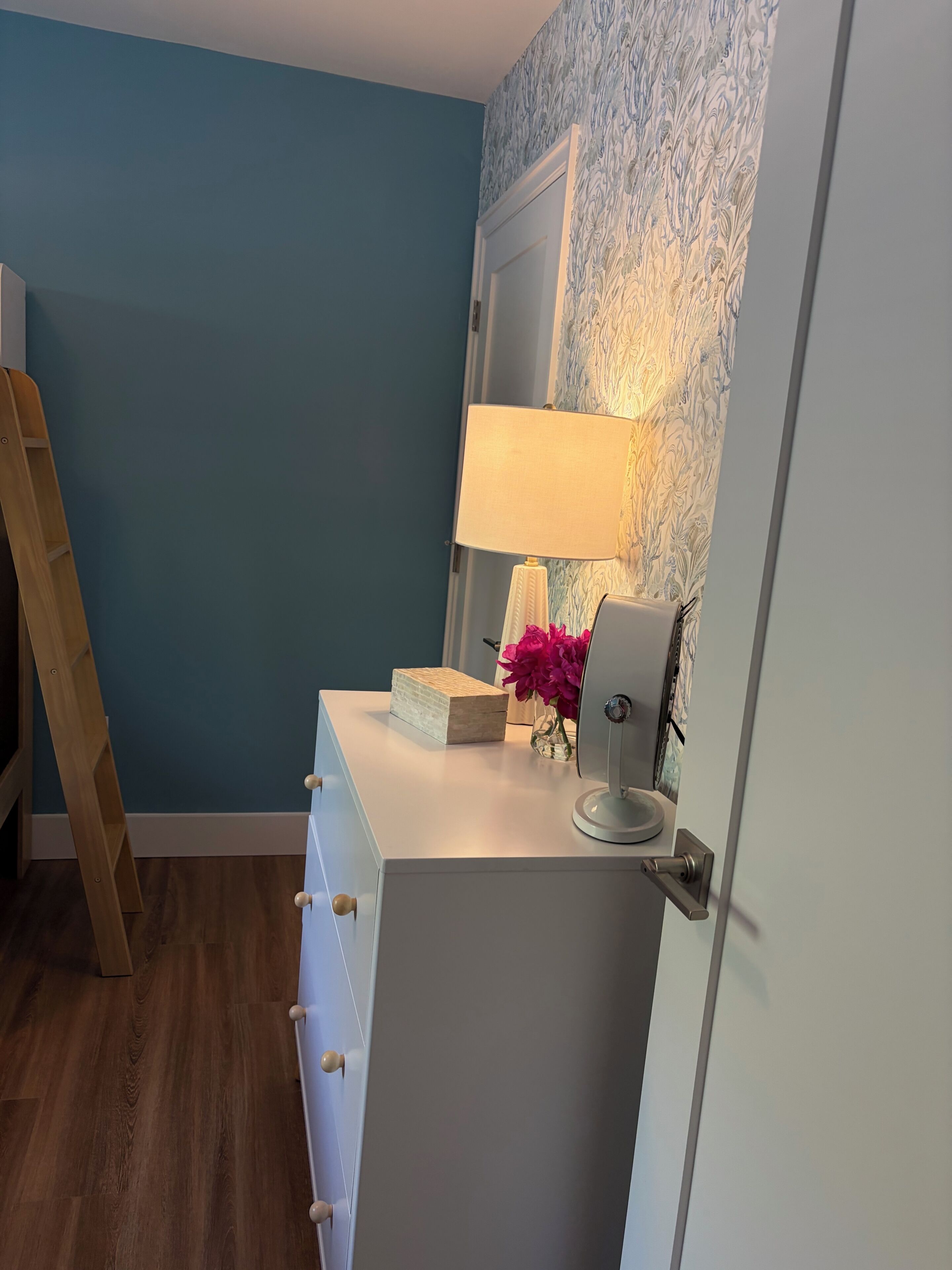 Ein Zimmer mit einer Holzleiter, einem weißen Schreibtisch mit Schubladen, einer Lampe, einer Vase mit Blumen und einem gemusterten Wandpapier.