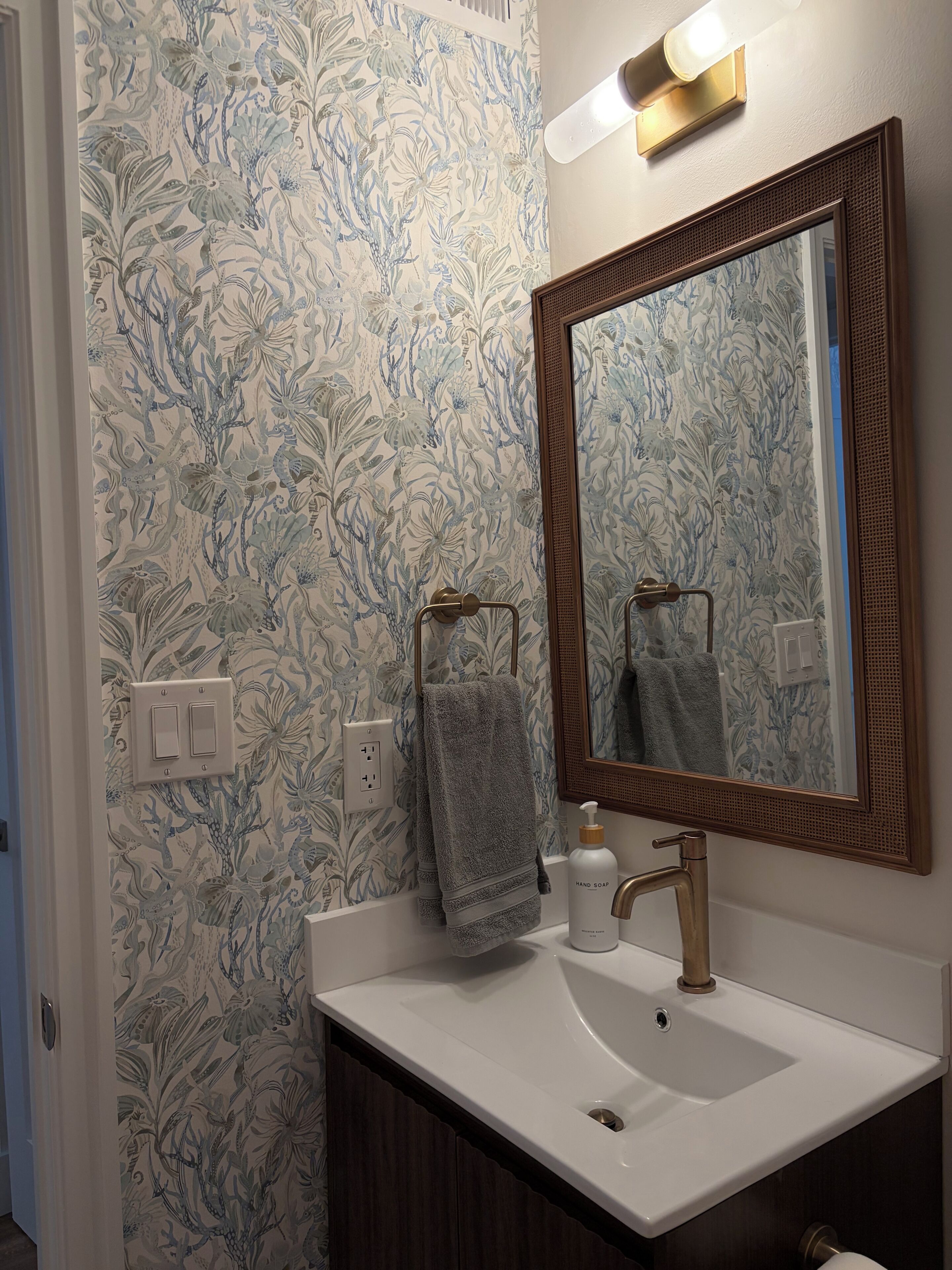 A bathroom with a floral wallpaper, a white sink, a mirror with a wooden frame, and a towel hanging on the wall.