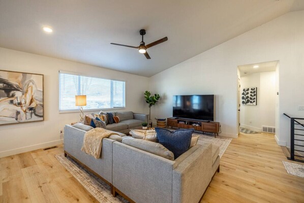 A spacious living room with a grey sectional sofa, a flat-screen TV, and a ceiling fan. - Meeting Place | Hot Tub, Theater Room, Mini Golf, Foosball (Sandy)