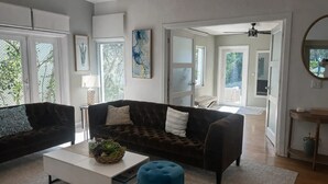 Ein großzügiges Wohnzimmer mit zwei dunklen Sofas, einem weißen Couchtisch und einem blauen Hocker. Große Fenster mit Jalousien, ein Deckenventilator und ein Spiegel an der Wand sind ebenfalls vorhanden.