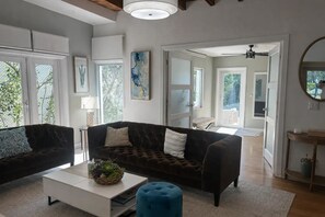 A spacious living room with two dark sofas, a white coffee table, and a blue ottoman. There are large windows with blinds, a ceiling fan, and a mirror on the wall. - Private Tropical Oasis w/Pool in Prime West Palm Beach (West Palm Beach)