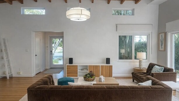 Ein großzügiges Wohnzimmer mit braunen Sofas, einem Couchtisch und einem Beistelltisch mit Lampe. Es gibt einen weißen Konsolentisch mit Lautsprechern und eine Tür zu einem anderen Raum.
