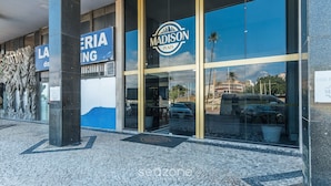 L’entrée de l’immeuble Madison Plaza, avec ses grandes fenêtres en verre qui reflètent les palmiers et les voitures.
