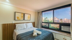 Une chambre d’hôtel comprenant un lit, deux œuvres d’art encadrées, une grande fenêtre donnant sur la ville et un appareil de climatisation.