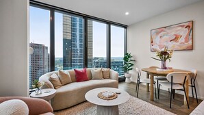 Una sala de estar moderna con sofá, mesa de centro y zona de comedor, con ventanas amplias y vista a la ciudad.