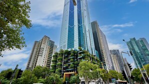 Un edificio de gran altura con fachada de vidrio y varios balcones, rodeado de otros edificios altos y árboles.
