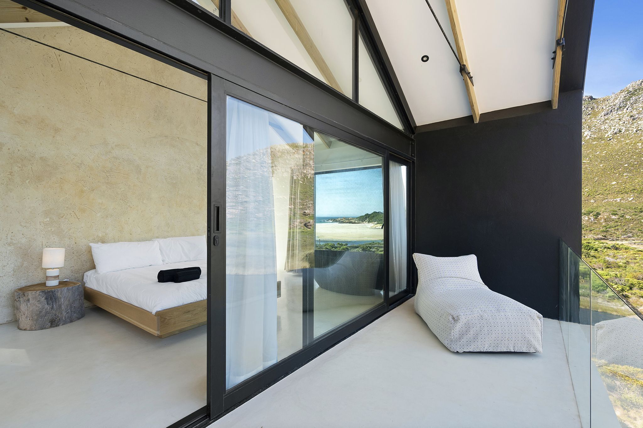 A modern bedroom with a large sliding glass door leading to a balcony, a bed with white bedding, and a small bedside table with a lamp.