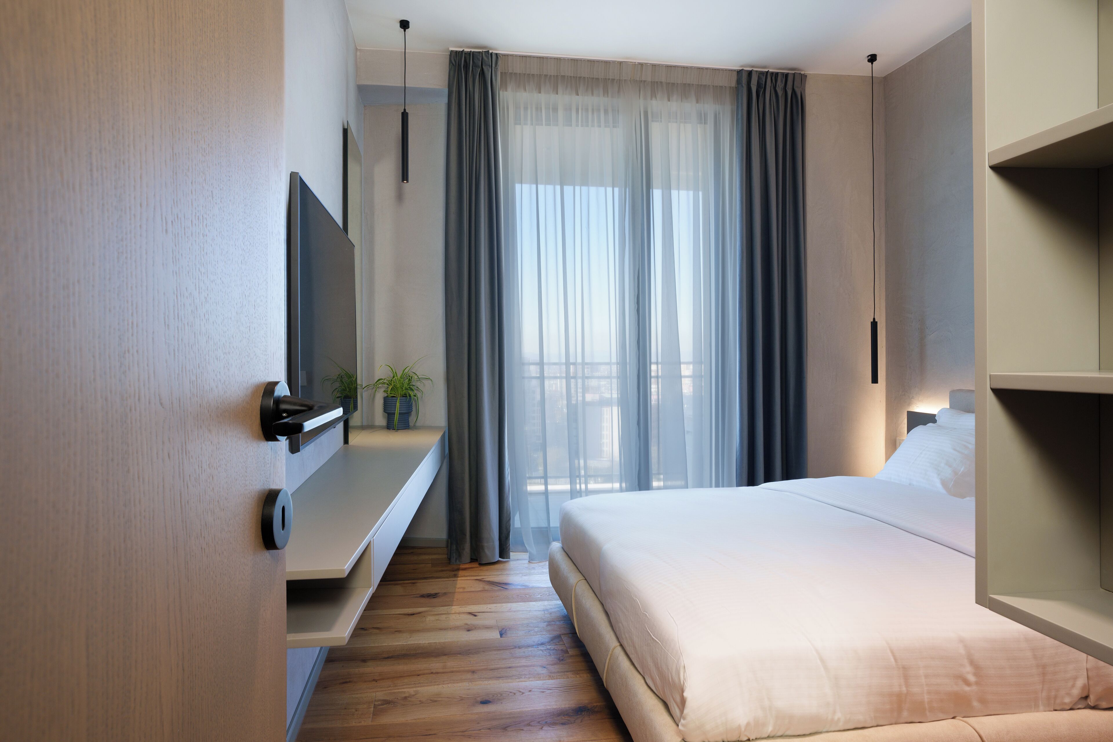 A modern hotel room with a bed, a TV mounted on the wall, a small table with a plant, and a balcony with curtains.