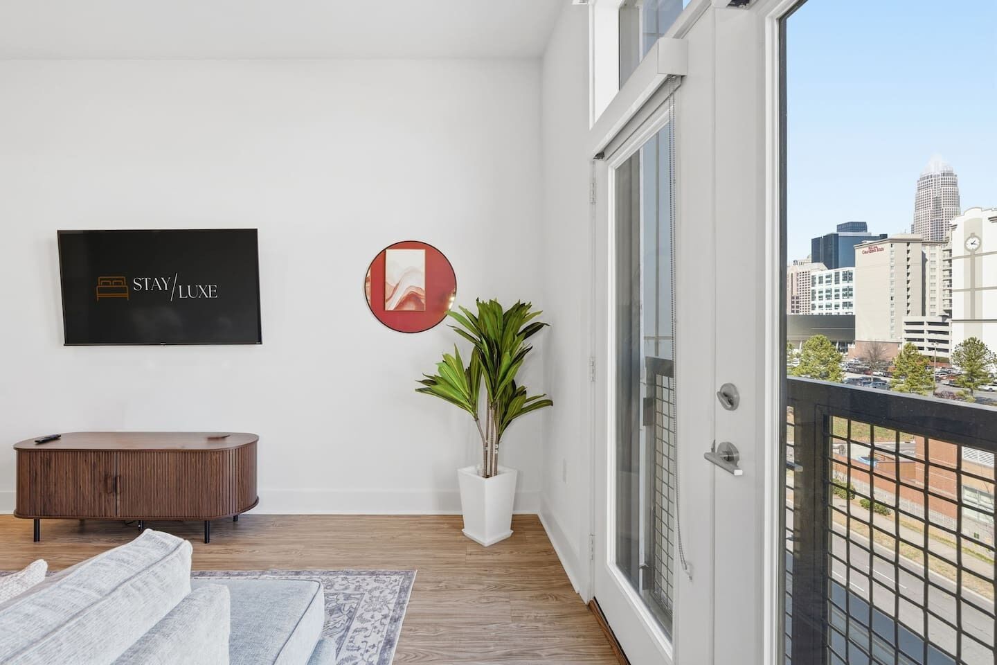 A modern hotel room with a large window, a plant, a framed artwork, a TV, and a wooden console.
