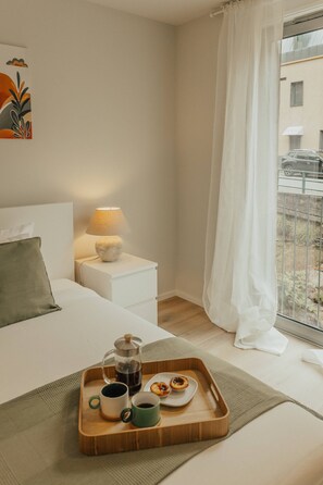 Habitació d'hotel amb un llit, una taula de nit amb una làmpada, una finestra que mostra un paisatge exterior, i una safata amb una cafetera, tasses i pastissos a terra.