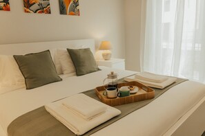 Una cama bien hecha con almohadas, una bandeja con cafetera, tazas y un pastelito, y una lámpara en una mesita de noche.
