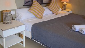 Ein ordentlich bezogenes Bett mit gestreiften Kissen, einer grauen Decke und einem aufgerollten Handtuch. Zwei Nachttische mit Lampen und ein Kunstwerk über dem Bett sind zu sehen.