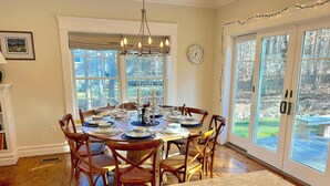 Une salle à manger avec une table en marbre, des chaises en bois et un lustre. Il y a des fenêtres avec des rideaux et une vue sur l’extérieur.