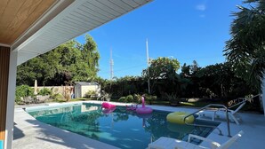 Una piscina cristalina con sillas de descanso y juguetes inflables, rodeada de follaje y un cielo despejado.