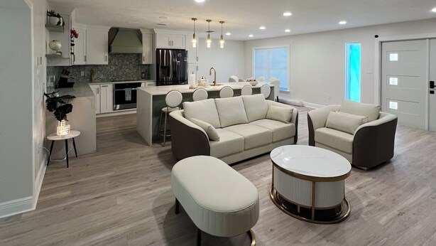 Una sala de estar moderna con sofá, reposapiés y mesa de centro, una cocina al fondo y un comedor con sillas.