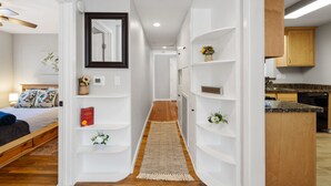 A narrow hallway with a wooden floor leading to a bedroom with a bed and a nightstand. Shelves line the walls, and a small rug is on the floor. - Red Door Cottage | 1/2 Mile to Whiskey Row (Prescott)